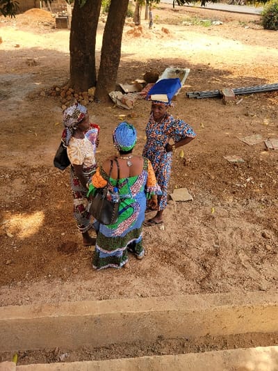 The image depicts an outdoor scene in Ebenebe, Nigeria, featuring three adult women conversing on a sandy-dirt ground under bright sunlight.
In the foreground, a light-colored concrete ledge or steps are visible. The midground features the three women standing in a loose semi-circle. The woman on the left wears a light-colored patterned dress and headwrap, with a black shoulder bag slung over her left shoulder. Her left profile is presented. The woman in the center has her back to the camera, dressed in a vibrant patterned gown with blue, green, and orange motifs, and a blue headwrap. A black shoulder bag hangs over her right shoulder. The woman on the right wears a blue and orange patterned dress with a matching headwrap. She carries a blue, fabric-covered rectangular object on her head. Her right hand rests on her hip, and she faces the other two women.
The ground is predominantly sandy-brown dirt, showing scattered debris including pieces of cardboard, several lengths of metal piping, and fragments of bricks or concrete blocks. Two thick tree trunks rise in the background behind the women. Further in the background, more open dirt ground, sparse vegetation, and indistinct structures or mounds are visible under clear conditions. Shadows indicate a high sun angle.