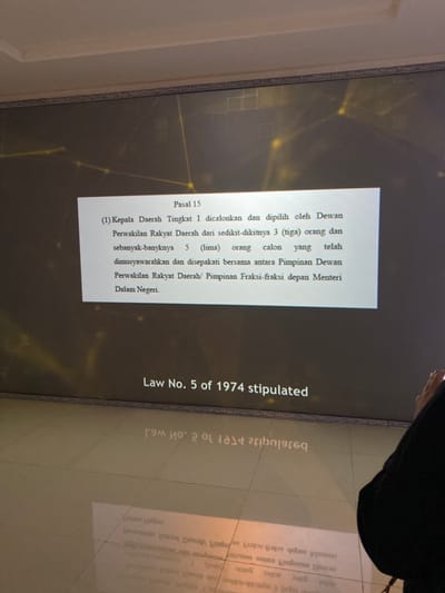 The image shows a screen with a text written in Indonesian. The screen is positioned on a wall in a room with light brown tiled floors. The text on the screen is "Pasal 15 (1) Kepala Daerah Tingkat I dicalonkan dan dipilih oleh Dewan Perwakilan Rakyat Daerah dari sedikit-dikitnya 3 (tiga) orang dan sebanyak-banyaknya 5 (lima) orang calon yang telah dimusyawarahkan dan disepakati bersama antara Pimpinan Dewan Perwakilan Rakyat Daerah/ Pimpinan Fraksi-fraksi depan Menteri Dalam Negeri. Law No. 5 of 1974 stipulated." The screen is reflecting onto the floor. A person wearing black clothing is standing to the right of the screen and is looking at the screen. This appears to be a scene from a room in an office building located at the coordinates -7.8 latitude and 110.407 longitude in Banguntapan, Indonesia.  The person is likely studying the text on the screen.  The room is well lit with the white light coming from a source unseen in the photo.