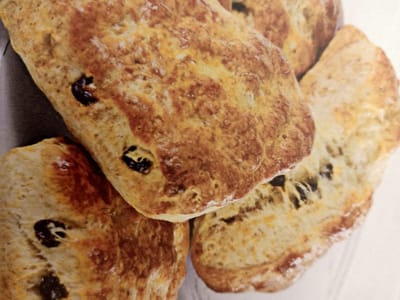 The image displays a close-up, overhead view of several freshly baked raisin scones. The scones are golden brown with darker, caramelized edges and visible raisins baked into the dough. They are arranged in a slightly overlapping cluster on a white surface. The lighting is bright, highlighting the texture and details of the baked goods. The environment is neutral, suggesting a food preparation or presentation setting. The image does not contain any elements that can be used to determine its location in Rumaithiya, Kuwait.