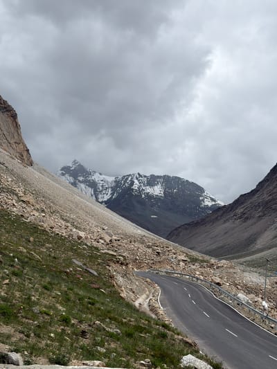A winding asphalt road curves through a mountainous valley in Darcha, India, under a cloudy sky. The road has white dashed lines marking the lanes and a metal guardrail on the right side. Steep, rocky slopes flank the road, with patches of green grass and scattered boulders. In the distance, snow-capped mountains rise dramatically against the overcast sky. The scene is devoid of people, with the landscape dominating the view.