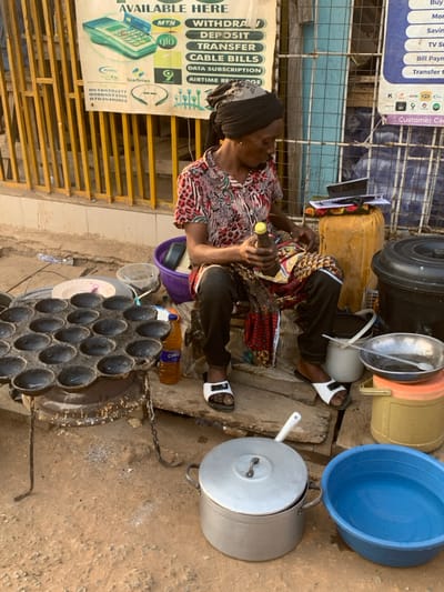 A woman sits preparing street food, likely "Puff-puff," using a specialized pan with multiple hemispherical molds over a stove. Various cooking utensils, including pots and bowls, surround her. She's situated beside a metal structure, with a sign promoting mobile banking services, like withdrawals, deposits, and data subscriptions.