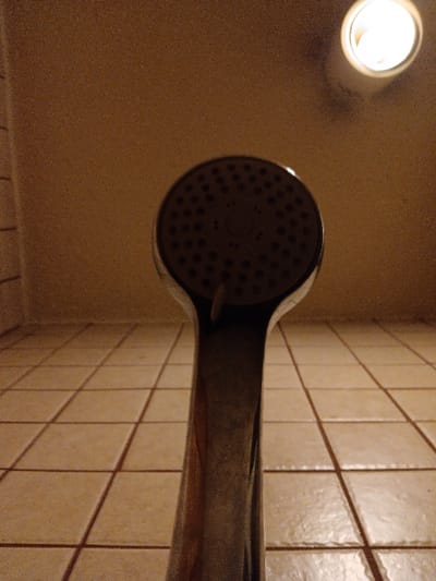 A showerhead with a chrome finish is positioned above a tiled floor. The showerhead is mounted to the wall, with a small white object hanging from it. The floor is beige and cream, tiled in a square pattern. The room is dimly lit, with a single light fixture visible in the upper right corner of the image. This shower is located in a bathroom in Riyadh, Saudi Arabia. The coordinates (latitude: 24.712, longitude: 46.696) place this shower in the Al-Malaz district of Riyadh.