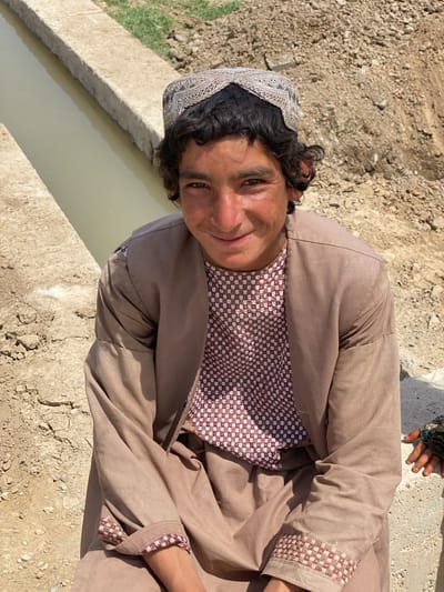 A young man with curly black hair and a decorative hat is sitting on the edge of a concrete irrigation canal in Ashoqah, Afghanistan. He is wearing a patterned brown and white shirt underneath a long-sleeved brown robe with patterned cuffs. His hands are resting in his lap. A small portion of another person's hand is visible on the right edge of the image. The canal contains murky water, and the surrounding environment is dry and earthen, with some green vegetation visible in the background.