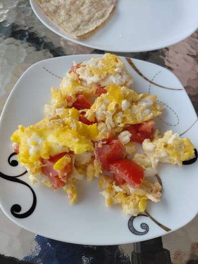 A close-up shot shows a white plate filled with scrambled eggs and chopped tomatoes. The eggs are fluffy and yellow, mixed with bright red pieces of tomato. The plate has a decorative border with thin gold lines and black swirl patterns. In the background, slightly out of focus, is another white plate with what appears to be a folded tortilla or flatbread. The table surface is reflective, showing a glass-like texture. The image does not provide any information about the location in Magnitogorsk, Russia, nor does it depict any people or actions.