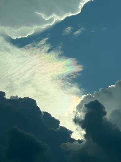 The image shows a sky filled with dramatic clouds, with a band of iridescence visible in the upper left quadrant. The clouds are dark and voluminous in the foreground, with lighter, wispy clouds in the background. The sky is a deep blue, indicating daytime. There are no people or structures visible in the image, and the scene appears to be a natural phenomenon occurring in the sky. The location is Tambon Don Sak, Thailand, but this context is not visually evident from the image itself.