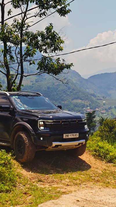 The image depicts a black Ford Raptor SUV parked on a dirt path with grassy surroundings, likely in a rural or mountainous area. The license plate reads "29K-179.83". A tree is positioned to the left of the vehicle. In the background, mountains and a distant village are visible, suggesting a location within the Hoàng Su Phì Commune in Vietnam. Power lines are strung across the upper right portion of the sky. The dirt path transitions into a paved road in the foreground.
