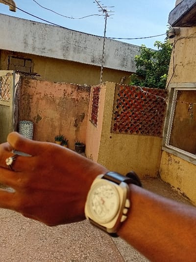 The scene, set in Sharada, Nigeria, depicts an outdoor environment under a clear blue sky. In the immediate foreground, a dark-skinned human left forearm and hand are prominently featured, presenting a first-person perspective. A square-faced wristwatch with a light-colored dial, a dark leather strap, and three visible push-buttons on the right side of the case is worn on the wrist. A silver-toned ring with a white stone is present on the ring finger. The ground surface directly beneath the hand is a light-colored, mottled concrete.
In the midground, an aged, light orange-pink plaster wall exhibiting weathering and dark stains extends horizontally. Sections of this wall are integrated with reddish-brown, circular-perforated brickwork acting as decorative screens. To the left of this wall, a white or light-colored cylindrical object with a patterned surface is visible. Small potted plants are discernible on the ground behind the orange wall. To the right, a section of a light yellow-brown building wall is visible, featuring a rectangular window frame.
The background shows the weathered, flat concrete roofline of a building. A tall metallic pole supporting a Yagi-Uda television antenna extends upward from behind this structure, silhouetted against the clear blue sky. Green tree foliage is visible to the right of the antenna. Dark-colored utility lines traverse the upper portion of the sky.