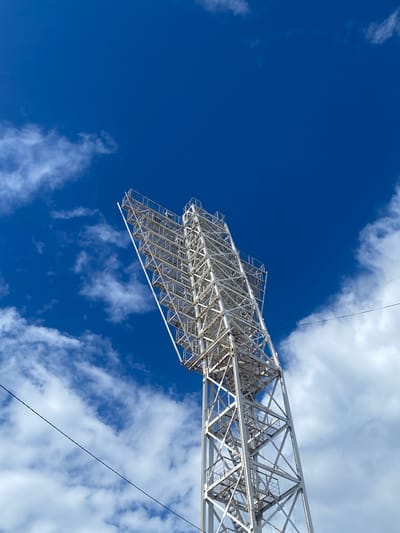 This media file captures a low-angle view of a large, metal lattice structure against a bright blue sky with scattered white clouds. The structure, which appears to be a lighting rig or part of a stadium grandstand, is made of white metal beams forming a complex geometric pattern. The perspective emphasizes the scale and height of the object, with the ground not visible. A thin black wire stretches diagonally across the frame.
The sky is a deep, vibrant blue, suggesting a clear day, possibly late morning or early afternoon, given the brightness. The clouds are wispy and white, indicating fair weather. There are no people visible in the frame. The overall impression is one of architectural grandeur and an expansive, open environment. The location context of Заволжье, Russia, is noted, but no specific geographical landmarks are identifiable within the image itself. The image is devoid of any visible text. The scene is neutral in emotional tone, focusing on the visual details of the structure and the sky.