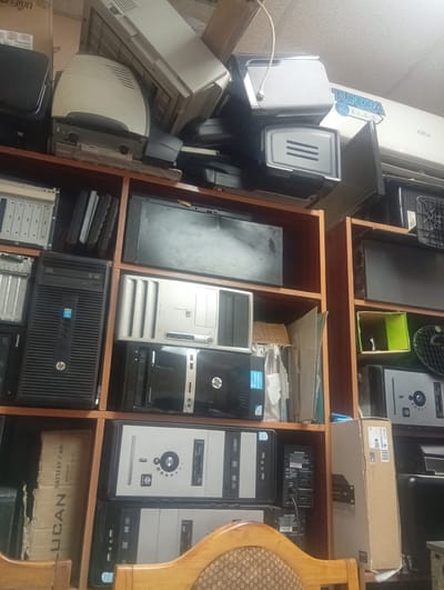 This media file depicts a cluttered storage area filled with various electronic equipment, primarily computer components. The scene is densely packed with shelves overflowing with old computer towers, monitors, power supplies, and what appear to be server racks. Some of the items are stacked haphazardly, indicating a disorganized storage environment. Visible text on the equipment and boxes includes brand names like HP, Compaq, and UniFi, as well as cautionary labels and specifications like "BATTERY 7 AH". The lighting suggests an indoor setting, possibly a workshop or a used electronics store, with fluorescent lighting from above. No people are visible in the image, and the overall impression is one of disuse and obsolescence, with the collection of older technology hinting at a place that deals with e-waste or vintage computer parts. The presence of a "TUNDRA" unit, possibly an air conditioner, on the upper shelf adds to the variety of items stored. The image captures a snapshot of a functional space that has become overwhelmed with its inventory.
