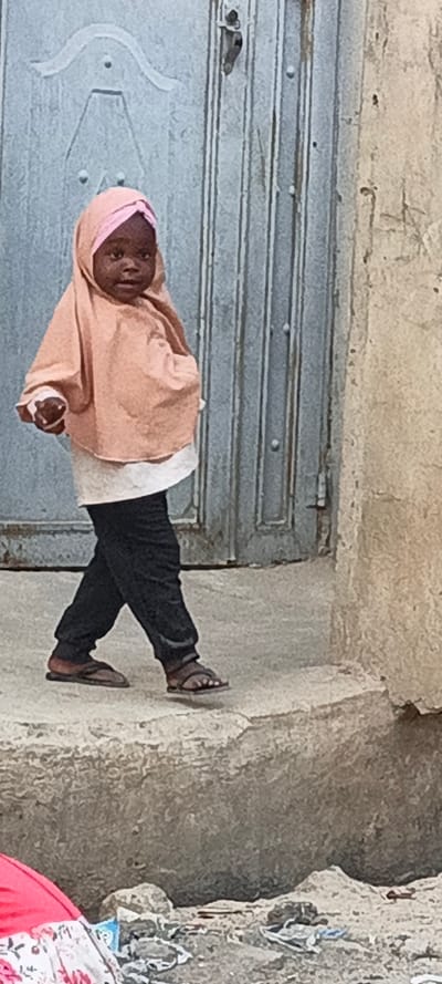 This vertical full-shot image captures a young child walking on a concrete surface next to a building in what appears to be an everyday setting in Rangaza, Nigeria. The child, with dark skin, is the central subject and is looking directly towards the viewer with a neutral, slightly serious expression.
The child is dressed in a light pink hijab or headscarf, a matching light pink long-sleeved top, a white undershirt, dark trousers, and dark sandals. Their left hand holds a small, light-colored object, possibly a piece of cloth or a small item.
The background consists of a weathered, grey metal door with visible vertical ridges and some ornamental detailing towards the top. To the right of the door is a rough, textured wall, light brown or tan in color, showing signs of wear. The child is walking on a raised concrete step or platform. In the bottom left corner, a lower ground level is visible, containing some indistinguishable debris and a crumpled piece of red and white patterned fabric.
The image appears to be taken during daylight hours, with soft lighting suggesting either an overcast day or a time when the sun is not directly overhead, as there are no harsh shadows. The scene evokes a candid moment of daily life in a residential or street environment. No visible text is present in the image.