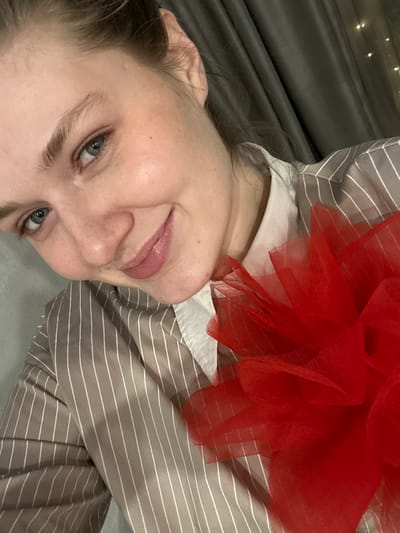 The image displays a female subject indoors in Borovoye Matyushino, Russia. The subject, an adult with light skin, blue eyes, and light brown eyebrows, is depicted from the upper torso upwards, angled slightly towards the camera. Her hair is pulled back, exposing her right ear. She is smiling, looking directly at the viewer, with slightly parted lips enhanced with a light pink gloss. She wears a light brown or taupe button-up shirt featuring thin white vertical stripes, with a white collar visible beneath. A substantial, multi-layered object constructed from bright red tulle or similar sheer fabric rests on her right shoulder and chest area. The immediate background consists of a solid light gray wall or surface on the left and dark gray curtains on the right. Blurred, faint lights, consistent with string lights, are discernible through or behind the curtains on the upper right side of the frame.