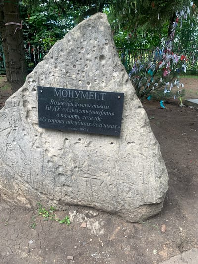 This media file shows a monument made of a large, rough-hewn stone situated outdoors. A black plaque is affixed to the stone with white text. The plaque reads: "МОНУМЕНТ Возведён коллективом НГДУ «Альметьевнефть» в память легенде «О сорока погибших девушках» июль 1997 г." This translates to "MONUMENT Erected by the staff of NGDU "Almetevneft" in memory of the legend "About forty fallen girls" July 1997."

The stone itself is textured and appears weathered, with numerous small indentations or pits on its surface. Behind the monument, a tree is visible, decorated with many colorful ribbons tied to its branches. These ribbons suggest a place of remembrance or prayer. The ground around the monument is dirt and appears somewhat dry and cracked in places, with some small green plants growing. The scene suggests an outdoor memorial site, possibly in a park or natural setting, dedicated to a historical event or group of people. The time of year appears to be summer, given the greenery and bright lighting, though the specific time of day is not clearly indicated. The overall mood is solemn due to the memorial nature of the monument, but the colorful ribbons add a touch of vibrancy.