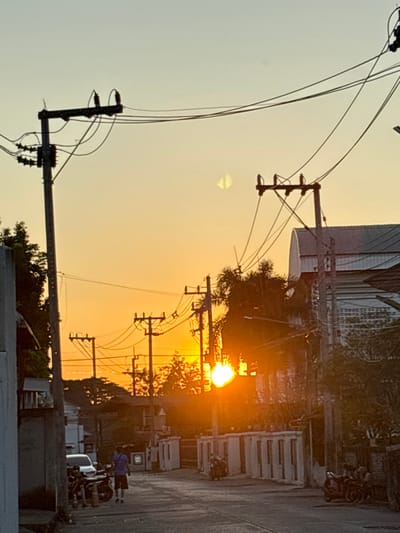 The scene depicts a street in Chiang Rai City Municipality, Thailand, at either sunrise or sunset. The sky is a clear gradient of light orange and yellow, with the sun visible as an intense, overexposed orange orb positioned in the mid-right, partially obscured by trees and utility poles. A translucent, semicircular lens flare artifact is present in the upper center-left sky.
Dominant features include numerous dark utility poles with multiple cross arms and insulators, supporting a dense network of black overhead electrical and communication wires that crisscross the upper portion of the frame. On the far left, a light-colored building segment frames the view.
A paved street, appearing dark due to backlighting, recedes into the background, curving slightly. On the left side of the street, a white sedan is parked, facing right, with two dark motorcycles parked alongside it. One individual, wearing a dark blue shirt and shorts, walks away from the camera on the left side of the street, towards the background. A second, less distinct individual is visible near the parked motorcycles.
Along the right side of the street, a series of uniform, light-colored fence posts or pillars define a property boundary. Further in the background on the right, additional dark motorcycles are parked. Beyond these, structures with dark roofs and walls are visible, including a prominent building with a light-colored, curved roof in the upper right background. Trees with dark foliage are interspersed throughout the scene, silhouetted against the bright sky.