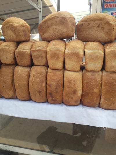 The image displays a close-up view of numerous loaves of bread arranged for sale, likely at a market stall or a small bakery in Ghafurov, Tajikistan. The bread loaves are mostly uniform in size and shape, featuring golden-brown, slightly textured crusts, characteristic of freshly baked bread.

The loaves are neatly organized. On the left side, several loaves, slightly larger and more irregularly shaped with a rounded top, are stacked or placed individually. To their right, a larger collection of bread, predominantly rectangular, is stacked in multiple rows on a white paper or cloth lining. This arrangement suggests a display within a container or on a counter. The bread appears to be a single type of traditional, rustic loaf.

In the blurred background on the upper left, a blue sign with partially visible Cyrillic text can be seen. The words "ФРУКТОВЫЙ" (Fruity), "КОКТЕЙЛ" (Cocktail), and "ЯХН" are discernible, indicating the presence of a fruit juice or cocktail stand nearby. The right side of the image shows a dark, reflective surface, possibly a glass display case or window, reflecting ambient light and indistinct shapes. The scene suggests a daytime setting within a commercial environment where food items are sold. No individuals are visible in the frame.