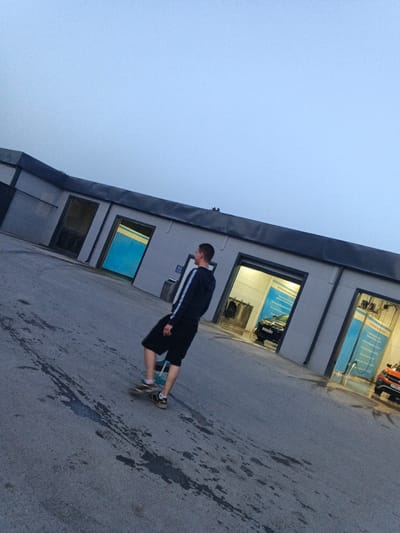 A man is seen from behind, skateboarding in front of a row of car wash bays in Spassk-Dal'nij, Russia. He is wearing black shorts and a dark blue hooded sweatshirt with white stripes on the sleeves. The car wash bays have open entrances revealing bright interiors and large blue banners. A black car is visible inside one of the bays, and an orange car is parked in front of another. The ground is paved and appears wet in places, with tire marks visible. The sky is overcast and light blue.