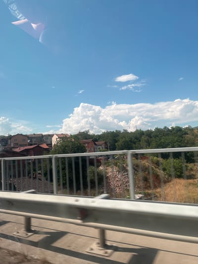 This image, taken in Dupnitsa, Bulgaria, captures a daytime scene viewed from what appears to be a bridge or elevated walkway, indicated by the presence of a white metal railing in the foreground and a concrete structure casting shadows. The perspective is from a slightly high angle looking down and across a residential area.
The main subjects are the houses and rooftops of a town, clustered together and set against a backdrop of lush green trees on a hillside. Some of the houses are multi-story buildings with varied architectural styles and roofing materials, primarily red and brown tiles. The area appears somewhat rugged, with piles of debris or construction materials visible on the slopes near the houses, suggesting ongoing development or a less manicured landscape.
Above the town and trees, a bright blue sky dominates the upper portion of the image, punctuated by large, fluffy white clouds, particularly on the left side. The clouds are billowy and suggest a fair weather day, likely a warm afternoon given the strong sunlight and shadows. The overall mood is peaceful and picturesque, with the natural elements of sky and greenery contrasting with the man-made structures. There are no visible people or specific activities, suggesting a general view of the town. The orientation of the image is also noteworthy as it is rotated 90 degrees clockwise.