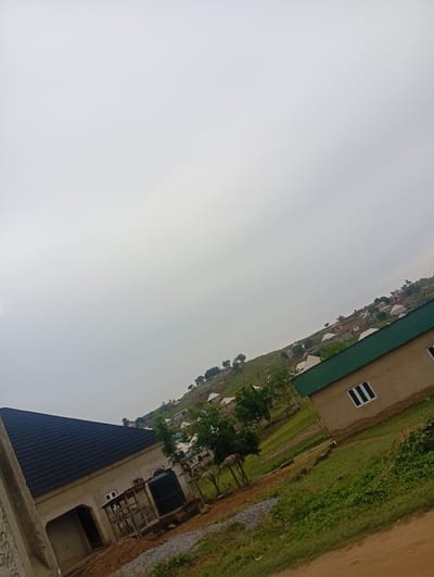 The image depicts a partially constructed residential area in Ashata, Nigeria, under an overcast sky. In the foreground, a building with a black, corrugated metal roof is visible. Adjacent to it, another structure with a light-colored facade and a green corrugated metal roof features a white-framed window with three vertical panes. A black cylindrical water tank stands next to a wooden support structure. The landscape is characterized by rolling hills dotted with houses of various sizes and colors, suggesting a developing community. Green vegetation, including trees and grass, covers the slopes. The ground in the immediate foreground is covered with dirt and gravel, with some patches of greenery. The overall impression is one of ongoing construction and settlement in a hilly, semi-rural environment.