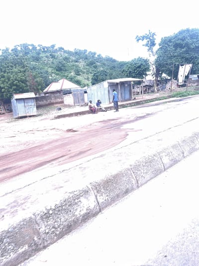 In Minda, Nigeria, a street scene unfolds with a dry, reddish-brown dirt road in the foreground, bordered by a concrete curb and gutter. Beyond the road, a collection of structures lines the path. To the left, a small, corrugated metal shed stands. Further back, several other corrugated metal buildings and a building with a red-tiled roof are visible, some appearing to be shops or residences. A few individuals are present: one person in a blue shirt and grey shorts stands near the metal shed, facing away from the camera. Two other people are seated on a low wall in front of some buildings, one wearing a patterned skirt. The background features a lush green, tree-covered hillside under a bright, overcast sky. The environment suggests a rural or semi-urban setting with simple infrastructure.