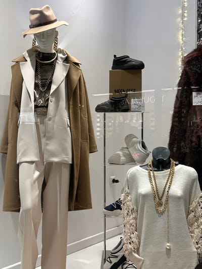 This media file captures an indoor retail display, likely from a clothing and accessories store in Koridallos, Greece. The image is oriented 90 degrees clockwise.
In the foreground, a headless mannequin is dressed in a smart ensemble. It wears a light tan fedora-style hat, a long, camel-colored overcoat, and a light-colored suit consisting of a jacket and trousers. Multiple gold-toned chain necklaces, some chunky and others more delicate, adorn its neck.
Behind a clear partition, another mannequin's torso is visible, wearing a white long-sleeved top or sweater with a decorative, textured trim around the neckline or shoulders. A long gold chain necklace with a pearl drop is draped over this mannequin.
Footwear is a significant part of the display. A pair of black boots is placed on a brown cardboard box labeled "MOON BOOT," with another pair of black boots visible nearby. Above these, a pair of light grey, chunky sneakers with a hint of orange detail is displayed. To the right, dark blue and white sneakers, along with parts of other white and black sneakers, are also arranged.
Visible text includes "MOON BOOT" on the cardboard box. Partially obscured price tags with handwritten details like "SNOW..." and "-50" are present, suggesting sale items. The setting appears to be well-lit by artificial indoor lighting, creating a clear view of the displayed fashion items. Reflections from the clear display surfaces are visible, adding to the multi-layered visual.