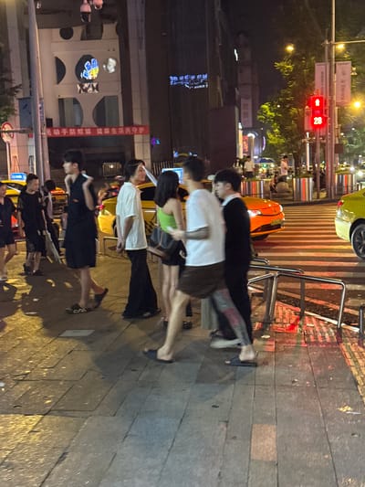 The image captures a street scene at night in Longta, China, likely a bustling urban area. The foreground features a pedestrian walkway paved with large, square tiles, some of which are wet and reflect the ambient city lights. A metal railing runs along the edge of the walkway, separating it from a crosswalk with prominent white stripes.
Several individuals are present on the walkway. To the left, a person in a black outfit is partially visible. Closer to the center, a young man in a black short-sleeved shirt and dark shorts walks past, his feet adorned with flip-flops. Next to him, a person in a white t-shirt and dark pants stands with their hands behind their back, their hair a light brown. Further to the right, a person in a lime green top and dark shorts is seen from the side. Another individual in a black long-sleeved shirt walks by on the far right.
In the background, yellow and orange vehicles, possibly taxis or private cars, are stopped at what appears to be a traffic light. The traffic light itself displays a red signal with the number "28" prominently displayed below it. Buildings with illuminated windows and signage rise on either side of the street. A banner with red Chinese characters hangs on the facade of one building. Trees with green foliage are visible, interspersed with streetlights that cast a warm glow. The overall atmosphere suggests a lively urban environment during nighttime hours.