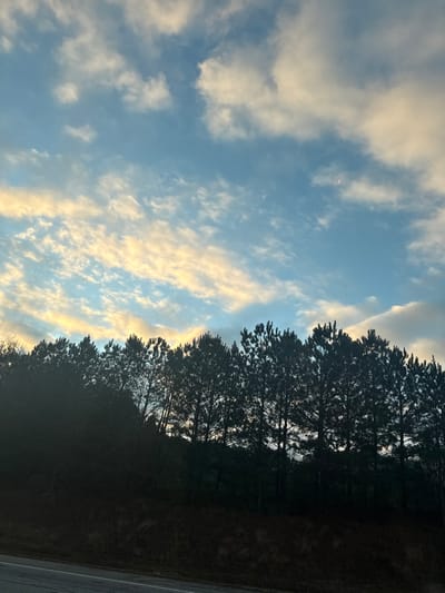 The image depicts an outdoor landscape scene within Lawrenceville, United States. The upper two-thirds of the frame consists of a partly cloudy sky, featuring patches of clear blue interspersed with white, grey, and horizontally elongated yellowish-orange clouds, suggesting either sunrise or sunset. A faint, reddish-orange crescent shape, possibly the moon or another celestial body, is visible in the upper right quadrant of the sky. The lower third of the image is dominated by a dark, silhouetted treeline composed primarily of coniferous trees, forming a dense horizontal band against the brighter sky. Below the treeline, a dark, vegetated slope descends towards the bottom of the frame. A dark, paved surface, likely a road or shoulder, is visible at the extreme bottom left corner, angling slightly upwards to the right. No individuals, vehicles, or specific human-made structures are discernible within the frame beyond the implied road. The lighting conditions indicate either early morning or late evening.