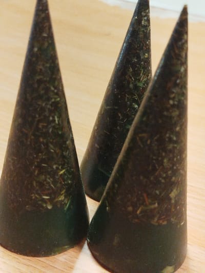 The image shows three dark green, cone-shaped objects arranged on a light-colored wooden surface. The cones appear to be made of a resinous material with visible inclusions of what looks like dried plant matter, possibly herbs or small leaves. They are positioned close to each other, with the tallest cone on the left and the others slightly behind. The background is out of focus, maintaining the focus on the cones. The light suggests an indoor setting, given the soft shadows and lack of direct sunlight. There is no indication that this picture was taken in Боровково, Russia.
