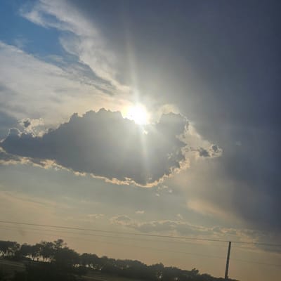 The image captures a dramatic sky over what appears to be a rural landscape, possibly in or near La Beltraneja, Spain. The sun is partially obscured by a large, dark cloud, emitting bright rays of light that pierce through the overcast conditions. Wispy, lighter clouds are scattered across the sky, with patches of blue visible in the upper left quadrant. In the lower portion of the image, a silhouetted treeline is visible, suggesting a natural or park-like setting. Several horizontal lines, likely power lines, stretch across the middle ground. A single wooden utility pole stands out on the right side of the frame. The overall impression is one of a dynamic sky, possibly indicative of an approaching storm or the aftermath of one, with the sun attempting to break through. There are no people or discernible actions taking place in the scene.