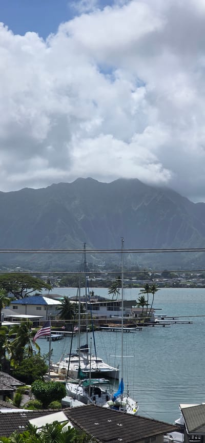 The image depicts a scenic view of Kaneohe, Hawaii, United States of America. In the foreground, several boats are docked in the clear blue water. Among them, a white catamaran stands out with its distinctive hull and multiple masts. Behind the boats, residential buildings with tiled roofs and palm trees line the shore. An American flag is prominently displayed on a flagpole near one of the houses. In the mid-ground, more buildings and vegetation are visible, suggesting a developed coastal area. The background is dominated by a majestic mountain range, partially shrouded in clouds, characteristic of Hawaii's lush volcanic landscape. The sky above is a mix of blue patches and fluffy white clouds, indicating a partly cloudy day. Several power lines stretch across the upper portion of the image, a common urban element. The overall scene conveys a tranquil, tropical atmosphere with a blend of natural beauty and human settlement.