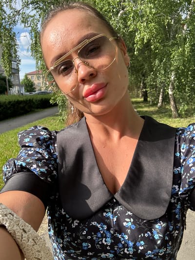 A woman with full lips and large gold-rimmed glasses is pictured in a close-up selfie. She is wearing a black dress with a blue floral print and a prominent black collar. The background suggests an outdoor urban setting, with trees and a sidewalk visible. The lighting indicates it is daytime, with sunlight casting shadows. A portion of a light-colored knitted sleeve is visible on the left side of the frame. This scene appears to be located in Yuzhnouralsk, Russia, based on the provided context.