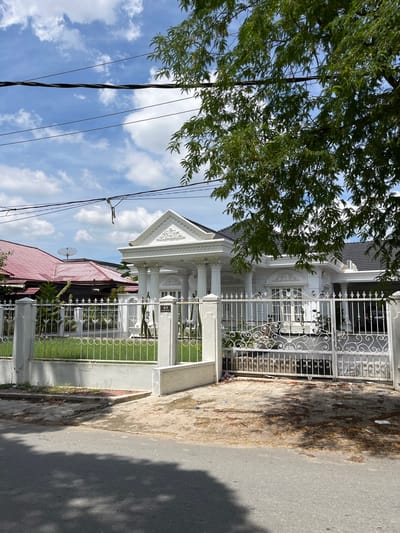 A bright, overcast day in Langsa, Indonesia, captures a view of a modern, white house with classical architectural elements, including columns and ornate trim. The house features a dark tiled roof and a large, decorative white metal gate. A white fence, also featuring decorative elements, surrounds the property. Adjacent to the modern house, another building with a red tiled roof and a satellite dish is visible. Power lines stretch across the top of the frame, and a large tree with green leaves partially obscures the view. The foreground shows a paved street and driveway littered with debris, including leaves and plastic waste. No people are visible in the image.