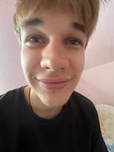 The image is a close-up, fisheye-lens selfie of a young Caucasian male with light brown hair and blue eyes. He is wearing a black t-shirt and appears to be indoors, with a pink wall in the background. There is no indication of the location in the city of Gari, Russia. The subject is looking directly at the camera with a slight smile. The lighting suggests it is daytime.