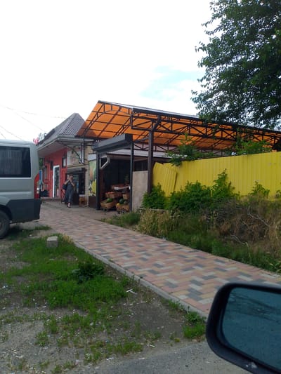 An outdoor street scene in Kochubeyevskoye, Russia, features a small market with a red-brick building and an adjacent open-air stall. A woman in a dark dress is standing at the entrance to the market, holding a phone to her ear. The market stall displays various fruits and vegetables, including watermelons and apples, in cardboard boxes. A silver van is parked to the left of the scene, and a yellow metal fence runs along the right side. A paved walkway with a colorful brick pattern leads past the market and stall. The area is lined with greenery, and a large tree is visible in the upper right corner. The sky is overcast with some light blue patches.