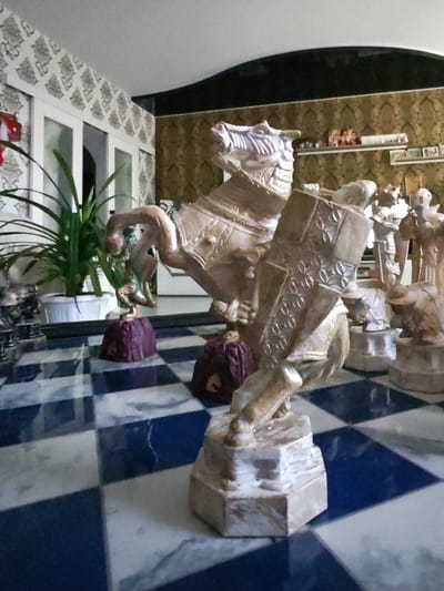 The image shows a close-up view of a chess set on a checkered board. The chess pieces are elaborate, carved from what appears to be a stone material, with detailed representations of horses and knights. The knight piece in the foreground is particularly detailed, with a shield adorned with a circular pattern. The background suggests an indoor setting with patterned wallpaper and shelves holding decorative items, possibly in a residential or display area. The floor is a reflective chessboard pattern, with alternating dark blue and white squares. The lighting is somewhat dim, casting soft shadows. The location is specified as Чонкаўскі сельскі Савет, Belarus.