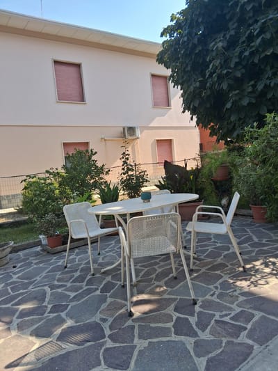 The image depicts a sunny day in the city of Colorno, Italy, in a residential backyard. A pale pink building with two windows that have red roller shutters is visible in the background. To the right of the building, a large tree with dark green leaves casts shadows on the scene. In the foreground, a patio is paved with irregularly shaped gray stones. A white oval table with four white perforated metal chairs is set up on the patio. The table has a small blue decorative object on it. Around the patio, there are various potted plants and a small garden area separated by a metal fence. The overall atmosphere is peaceful and suggests an outdoor seating area for relaxation or dining.