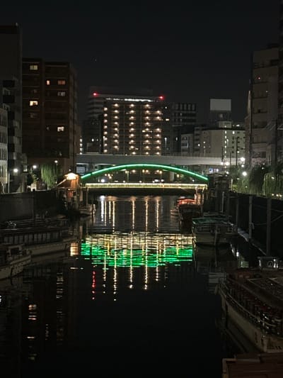 The image depicts a nighttime urban waterscape in Chūō, Japan, observed from a low vantage point looking upstream or across a canal. A prominent arched bridge, illuminated with vibrant green lights, spans the dark waterway in the mid-ground. The bridge's deck displays smaller, warm-toned lights and obscured vehicle activity.

On the left bank, a multi-story building with numerous warm-yellow illuminated windows rises against the dark sky. Below it, closer to the water, a smaller, dark structure with a light-colored roof and a single warm light is visible. Green, weeping foliage hangs over the water's edge. Several boats are moored along this bank, including a large, multi-deck vessel prominent in the left foreground.

On the right bank, additional multi-story buildings with illuminated windows are visible. Smaller boats are moored along this side, and similar green foliage extends over the water. Streetlights provide localized illumination.

The dark, calm water of the canal highly reflects the green bridge lights, creating a shimmering, distorted pattern, and also mirrors warmer lights from buildings and moored vessels.

The background consists of a dense urban skyline against a dark, starless sky. Numerous buildings display internal lighting, and some taller structures feature red anti-collision lights on their rooftops. A single, faint point of light is discernible in the upper center of the sky. No human figures are explicitly visible in the scene.