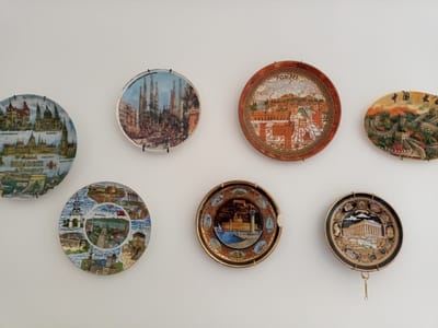 The image shows a collection of decorative plates displayed on a white wall. The plates are arranged in two rows.
In the top row, from left to right, there is a plate depicting buildings labeled "BUDAPEST Hungary," a plate with an image of the Sagrada Familia in Barcelona, Spain, a plate with a scene labeled "POMPEII," and a plate featuring the Great Wall of China.
In the bottom row, from left to right, there is a plate with various scenes and labels including "Alanya," "Side," "Konakli," "Belek," "Kemer," "Aspendos," and "Antalya," a plate with a lighthouse and harbor scene labeled "RHODES," and a plate depicting the Parthenon in Athens, Greece.
The plates are made of different materials, with some appearing to be ceramic and others possibly metal. They are all hanging on the wall, with visible hooks or wires supporting them. The lighting is even, suggesting an indoor setting with diffused light. There are no people visible in the image. The white wall serves as a neutral background, emphasizing the colorful designs on the plates.
The location of this collection of decorative plates is not specified in the image or the provided crops. Therefore, it is not possible to determine if it is located in the city of Be'er-Sheva, Israel.