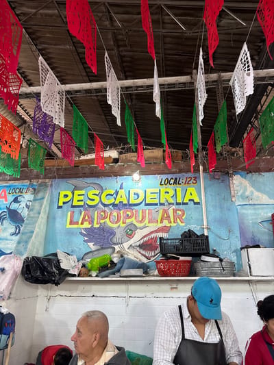 A vibrant Mexican market scene is shown. Colorful Papel Picado banners hang overhead. A fish stall, "Pescaderia La Popular LOCAL 123", features a whimsical shark mural. People are visible, including a vendor in a blue hat and apron and two customers, an older man and a child. The overall impression is one of a bustling and lively marketplace.