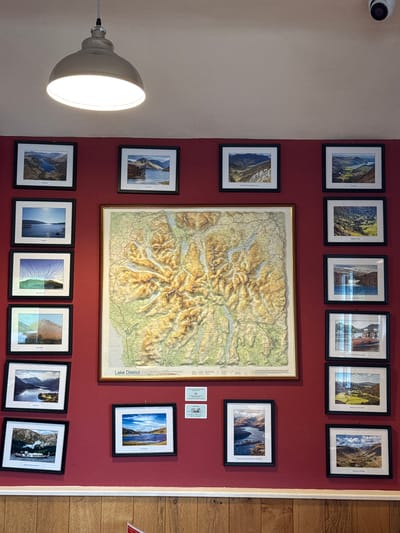 The image shows an interior wall of a room in Bowness-on-Windermere, United Kingdom, painted in a deep red hue, on which a collection of framed pictures and a framed map are hung. Above, a surveillance camera is mounted on a beige wall.

The pictures, framed in black, depict landscapes. A large, central framed map of the Lake District is prominently displayed. Below and to the side of the map are additional framed landscape pictures. There are twelve frames in total - six on the top row and six on the bottom row. On the right side of the map there are two small white cards with text.

A gray metal pendant light with a bright bulb is visible on the left side of the frame, obscuring part of the wall and some of the pictures. The floor on the right is made of light brown wood.