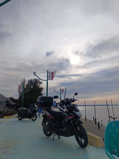 This image depicts a waterfront scene in Lancok, Indonesia, under a partly cloudy sky. In the foreground, two black motorcycles are parked on a light blue paved surface. The motorcycle on the right is closer to the viewer, showing its front wheel, part of the engine, and a black storage box on the rear. The motorcycle on the left is further back and appears to be a scooter.
Behind the motorcycles, a wooden pier extends into the water, with several vertical wooden posts visible along its edge. The water is calm and meets a distant shoreline under the diffused sunlight. To the left, a weathered, single-story building with a dark roof stands near the water's edge, surrounded by trees.
Two decorative lampposts with flag-like banners are also present, adding a touch of local character to the scene. The overall atmosphere is peaceful and suggests a casual, outdoor setting.