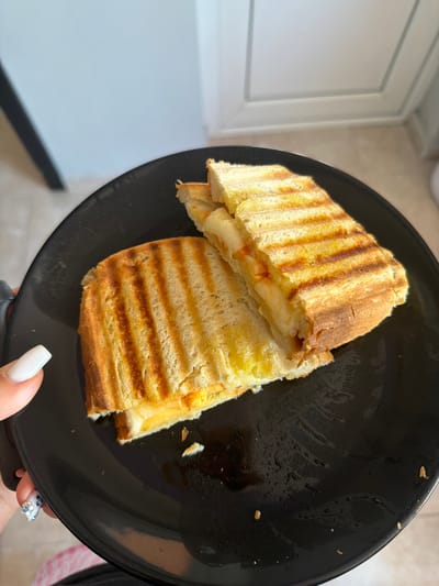 A close-up, eye-level shot shows two halves of a grilled cheese sandwich on a black plate. The sandwich is cut diagonally and shows melted cheese oozing out. The bread is golden brown with distinct grill marks. A person's hand with white nail polish is visible on the left side of the frame, reaching towards the plate. The plate has some moisture and crumbs on it. The background is out of focus, showing a light-colored wall and a doorway with white trim. The scene appears to be indoors, possibly in a kitchen or dining area. There is no visible indication of the city of Novi Iskar, Bulgaria.