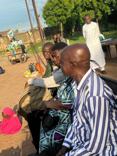 This outdoor scene, set in the city of Gbaza, Nigeria, depicts a gathering of individuals, primarily adult males, under natural daylight, likely during morning or late afternoon. The ground is unpaved reddish-brown dirt.

In the foreground on the right, an older, bald man with dark skin is seated, wearing a blue and white vertically striped long-sleeved shirt, looking to his left. Adjacent to him on the left, a person is partially visible, wearing a patterned garment in shades of blue, black, and purple. To their left, a man with dark skin is seated, wearing a grey long-sleeved shirt, with a light brown backpack resting on his lap. Further left, another man in a dark green shirt is also seated, looking leftward. On the far left of the foreground, a young child completely covered in vibrant pink fabric is seated on the dirt ground, facing downwards.

In the midground on the right, a man with dark skin stands behind a rustic wooden table, wearing a long white traditional garment and dark sandals. A green pen is visible in his shirt pocket. In the background on the left, a wooden vendor cart with two wheels is loaded with numerous plastic bottles containing various beverages. Several plastic bags in black, orange, and yellow are on the ground near the cart. Multiple rusty metal utility poles with overhead power lines are present in the left-midground and background. The backdrop includes a weathered concrete wall, distant buildings with corrugated roofs, and lush green trees under a light blue sky with scattered clouds.