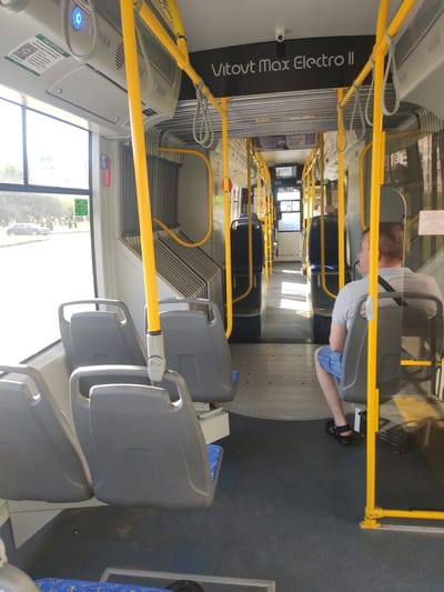 The image shows the interior of a bus. In the foreground, there are grey plastic seats with yellow metal handrails. Further down the bus, more seats are visible. A person is seated in one of the seats, wearing a grey t-shirt and blue shorts, and black sandals. They are facing away from the camera. The bus appears to be stationary.
On the side of the bus, text reads "Vitovt max Electro II". There are also some signs with Cyrillic text, one of which contains a QR code. One sign has the text "БЕЛАРУСБАНК". The lighting suggests it is daytime, and the sunlight streaming through the windows indicates clear weather. The scene is set inside a modern public transport vehicle, likely in Navapolatsk, Belarus, given the textual cues. The overall atmosphere is ordinary, depicting a typical scene inside a bus.