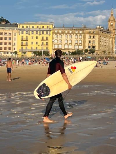 A surfer walks on a wet sandy beach towards the right, holding a light-colored surfboard. The surfer is wearing a dark wetsuit with maroon sleeves and is barefoot, with a pink ankle strap visible on their right ankle. The surfboard has a logo with three colored hexagons and text below it, and a black and purple fin area. In the background, numerous beachgoers are scattered across the sand, some sitting on towels and umbrellas, others standing. Behind the beachgoers, a promenade lines the shore, with several multi-story buildings in the background. The architecture of the buildings is characteristic of San Sebastián, Spain, featuring colorful facades and ornate detailing. The building on the right has a prominent clock tower. The sky is blue with scattered clouds. The lighting suggests it is late afternoon or early evening.