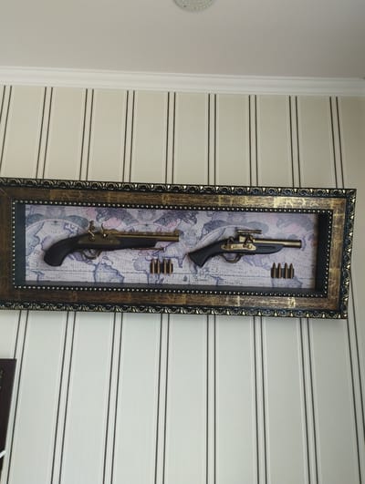 This is a medium shot, eye-level photograph of a framed display of two antique-style replica firearms and several bullets. The display is mounted on a wall with vertical striped wallpaper in off-white and thin black lines. The frame itself is ornate, with a dark brown base and gold decorative accents. The background within the frame is a sepia-toned map.
The firearm on the left is a single-barreled pistol with a dark brown grip and a golden barrel and hammer mechanism. To its right, and slightly lower, are four gold-colored bullets.
The firearm on the right is a rifle with a similar dark brown body and golden accents, featuring what appears to be a scope mounted on top. To its right, and slightly lower, are four more gold-colored bullets.
There are no people or animals visible in the image. The overall impression is of a decorative display intended to evoke a sense of history or adventure. There is no information in the image that allows for determination of its location in the city of Hrazdan, Armenia.