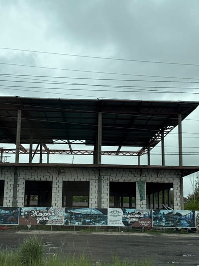 The image shows an unfinished building under construction in what appears to be Novosaratovka, Russia, based on the Cyrillic lettering on nearby advertising banners. The building has a metal roof structure, concrete supports, and walls constructed from white blocks with window openings. Power lines are visible in the sky. Adjacent to the building is a series of banners advertising "Women's Day" and "Family Day" discounts, indicating local business promotions. A road and some grass are in the foreground to the right of the building.