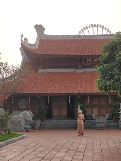 A full outdoor shot shows a woman standing on a brick-paved courtyard in front of a temple in the city of Hong Gai Ward, Vietnam. The woman faces the camera and looks down, holding a small device in her hands. She wears a tan dress, sneakers, and a pink visor. Behind her is a large, two-story temple with a red tile roof, wooden doors, and a Chinese inscription above the entrance. To the left of the temple is a stone statue of two horses, and to the right is a green tree. A Ferris wheel is visible in the distant background. The sky is overcast.