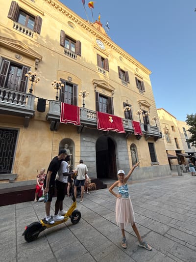 In Mataró, Spain, a scene unfolds in front of a tan-colored building with an ornate facade and multiple balconies. Two men are on an electric scooter in the foreground, with one appearing to be teaching the other to ride. A young girl in a blue and white patterned top and a light pink skirt strikes a pose with her arm extended, facing the camera. In the background, a woman sits on a bench, and several other people are visible walking and standing. A dog is also present in the mid-ground. The building has red banners hanging from its balconies, and a clock is visible near the roofline. The ground is paved with square tiles. The sky is clear blue, indicating daytime.