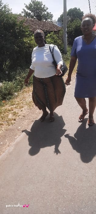Two adult females are observed walking on a paved pathway under direct sunlight in Homa Bay, Kenya. The woman on the left, facing slightly forward, wears a white short-sleeved top, a long brown and dark-patterned skirt, and dark sandals. She carries a black shoulder bag on her left shoulder, and her left hand is slightly extended forward. The woman on the right, partially visible, wears a dark blue short-sleeved dress and light-colored sandals. Both individuals cast distinct, elongated shadows on the pathway, indicating bright, high-angle sunlight. In the background, to the left, a building with a reddish-brown tiled roof is partially obscured by dense green foliage. Numerous trees and bushes line the background and right side. A utility pole is visible on the right side, behind the women. To the immediate left of the paved path is an unpaved strip of ground covered with sparse, dry vegetation.