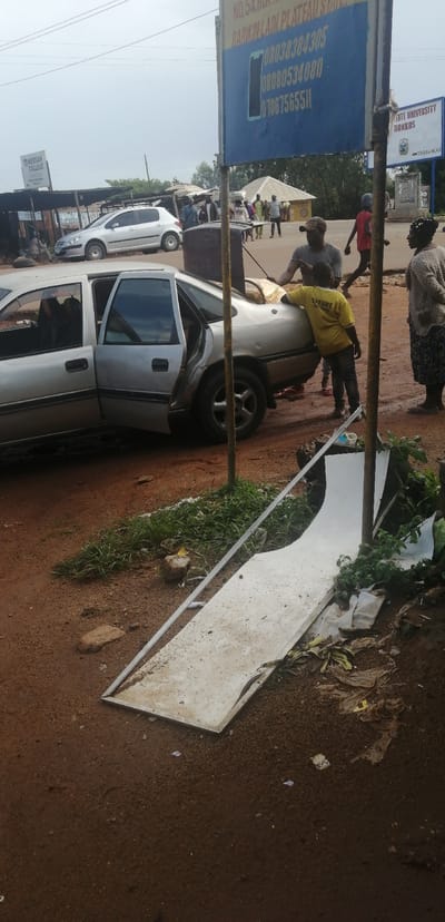 The image depicts a street scene in Kassa Market, Nigeria. In the foreground, a silver sedan with its doors and trunk open is parked on a dirt road. The trunk appears to be loaded with various items, and a young boy in a yellow shirt is leaning against the car. Two adults are visible near the car, one man with a cap and another woman in a patterned skirt and sweater. In the background, several market stalls with corrugated metal roofs are set up. A white hatchback is parked further down the road. A blue sign with white text and a phone icon is prominently displayed, listing phone numbers. Another sign, likely for a university, is visible on the right. The ground is covered in reddish-brown dirt and scattered debris, including what appears to be a large, flat white object with a metallic edge lying on the ground in the lower portion of the image, possibly a discarded sign or panel. Vegetation, including grass and some leafy plants, is growing along the side of the road. The sky is overcast.