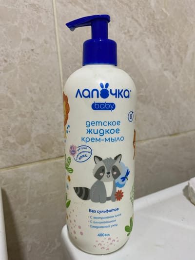 A white plastic bottle with a blue pump dispenser stands upright against a tiled wall. The bottle has a blue logo with bunny ears and the text "ЛАПОЧКА baby" printed in blue. Below this, "детское жидкое крем-мыло" is printed in blue, meaning "children's liquid cream soap." The bottle features colorful cartoon illustrations of animals, including a lion, a raccoon, and a duck, along with various plants and flowers. There is a small circular icon with a "0+" symbol. The text "Без сульфатов" (Without sulfates) is printed in blue, followed by bullet points indicating "с экстрактом алоэ" (with aloe extract), "с аллантоином" (with allantoin), and "ежедневный уход" (daily care). At the bottom, "400 мл" (400 ml) is written in blue. The background is a tiled wall with a beige and light brown marble pattern. The scene appears to be a bathroom. The location of Dobrovske rural settlement, Ukraine is not visually discernible from the image.